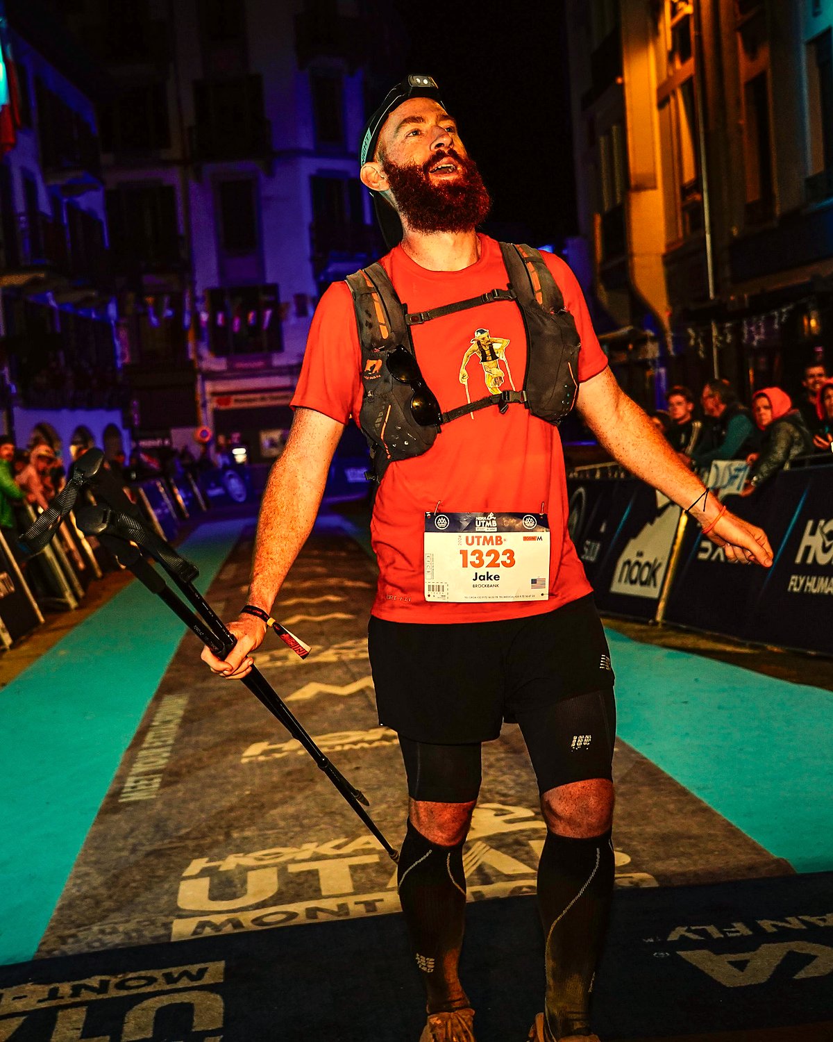 Jake Brockbank coming down the UTMB 2024 finish chute at night with his arms out and poles in hand